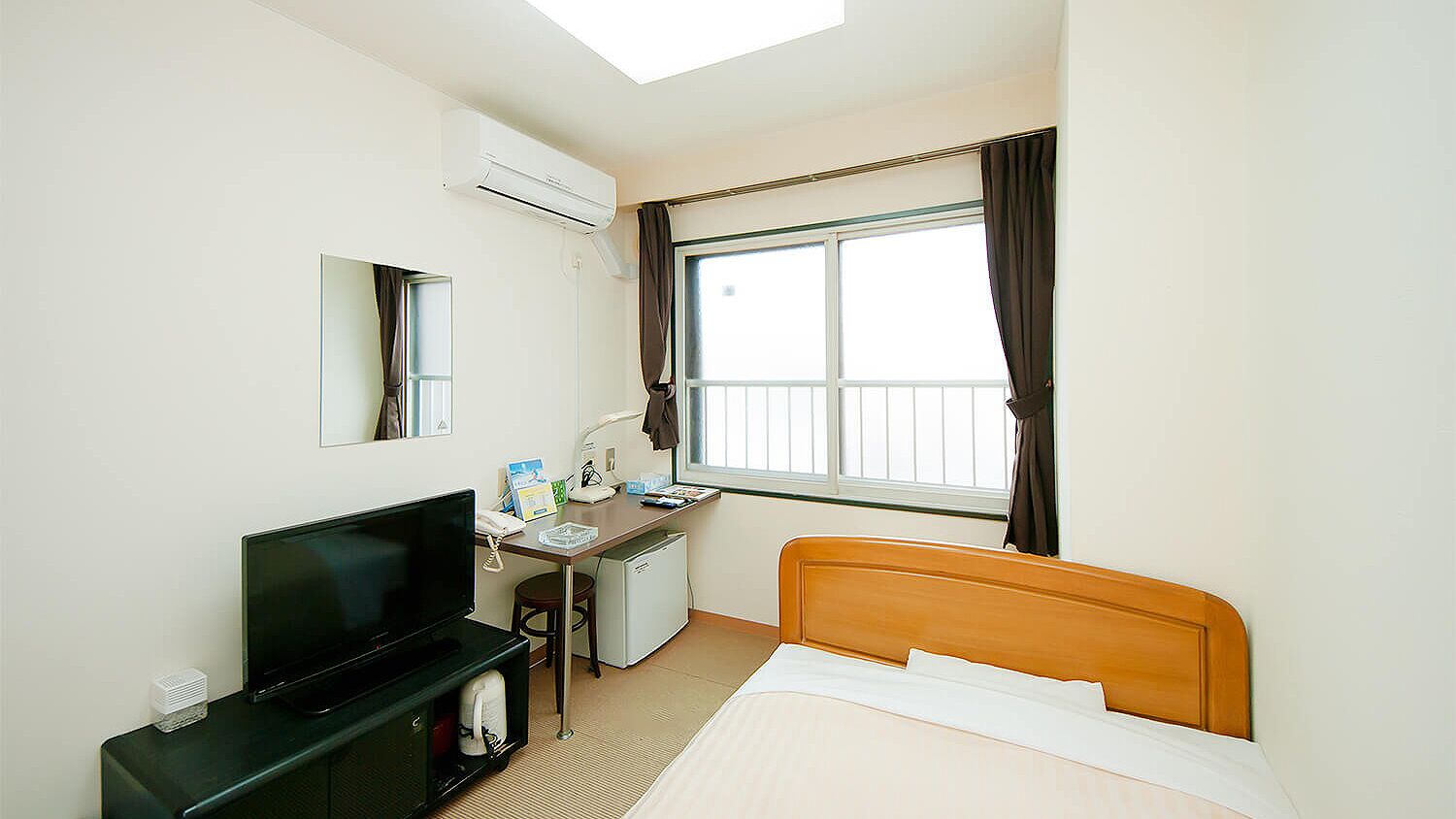 Habitación de hotel con cama, escritorio con computadora, televisor, refrigerador pequeño y ventana con cortinas.