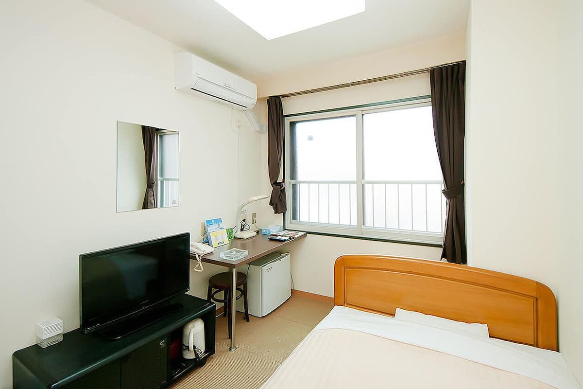 A hotel room with a bed, a desk with a computer, a television, a small refrigerator, and a window with curtains.