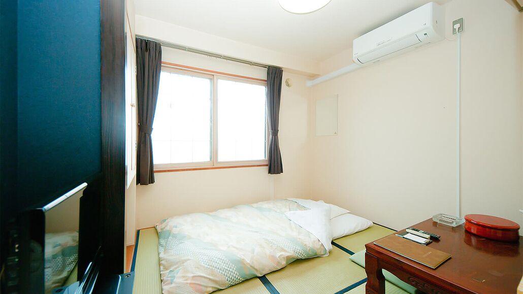 Una cama individual con una manta, una mesita de noche con un libro y un recipiente rojo, una ventana con cortinas y un aire acondicionado instalado en la pared.