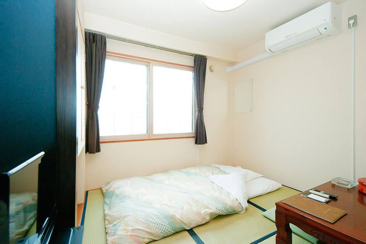 A single bed with a blanket, a bedside table with a book and a red container, a window with curtains, and a wall-mounted air conditioner.