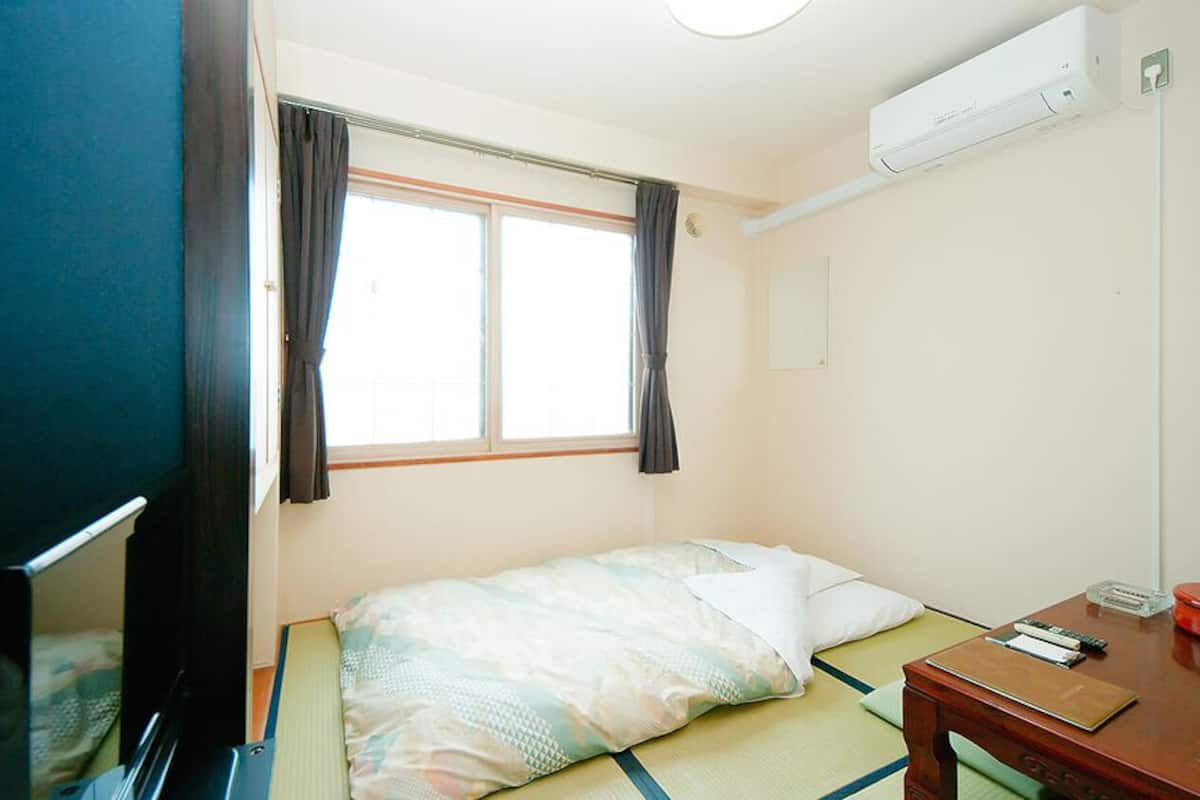 Una cama individual con una manta, una mesita de noche con un libro y un recipiente rojo, una ventana con cortinas y un aire acondicionado instalado en la pared.