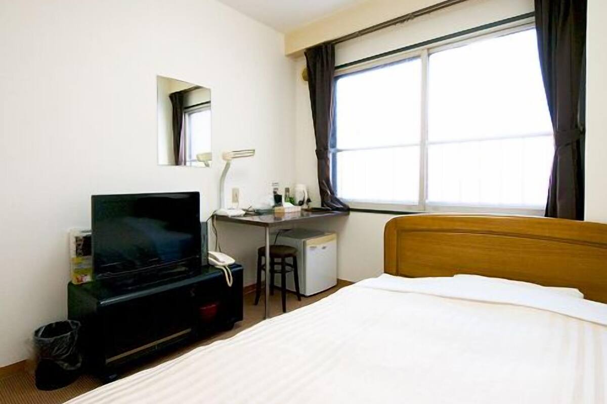 A hotel room with a bed, a desk with a computer, a TV, and a window with curtains.