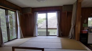 A room with a tatami mat bed, sliding glass doors, a small kitchenette, and a view of greenery outside. - K2 Cottage (Hachijo)
