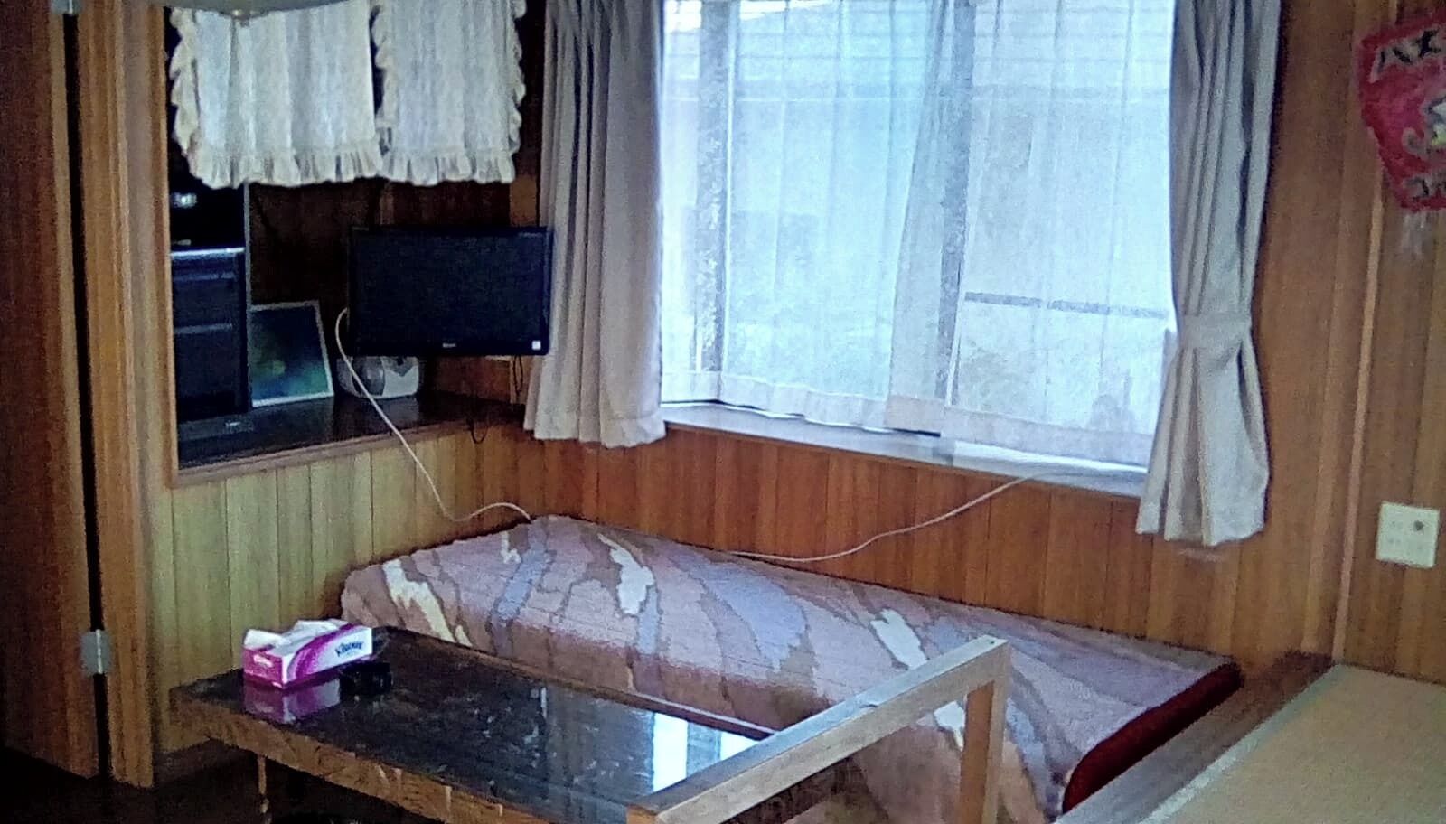 A room with a bed, a small table, a window with curtains, and a television on a shelf.