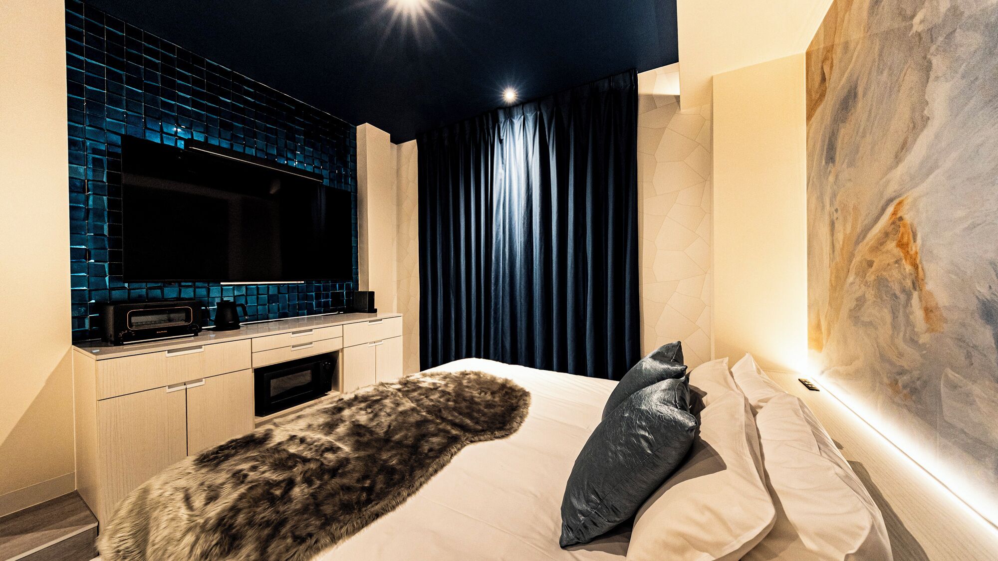 Quarto de hotel moderno com uma cama grande, televisão de tela plana, cozinha compacta e um quadro abstrato de grandes dimensões na parede.