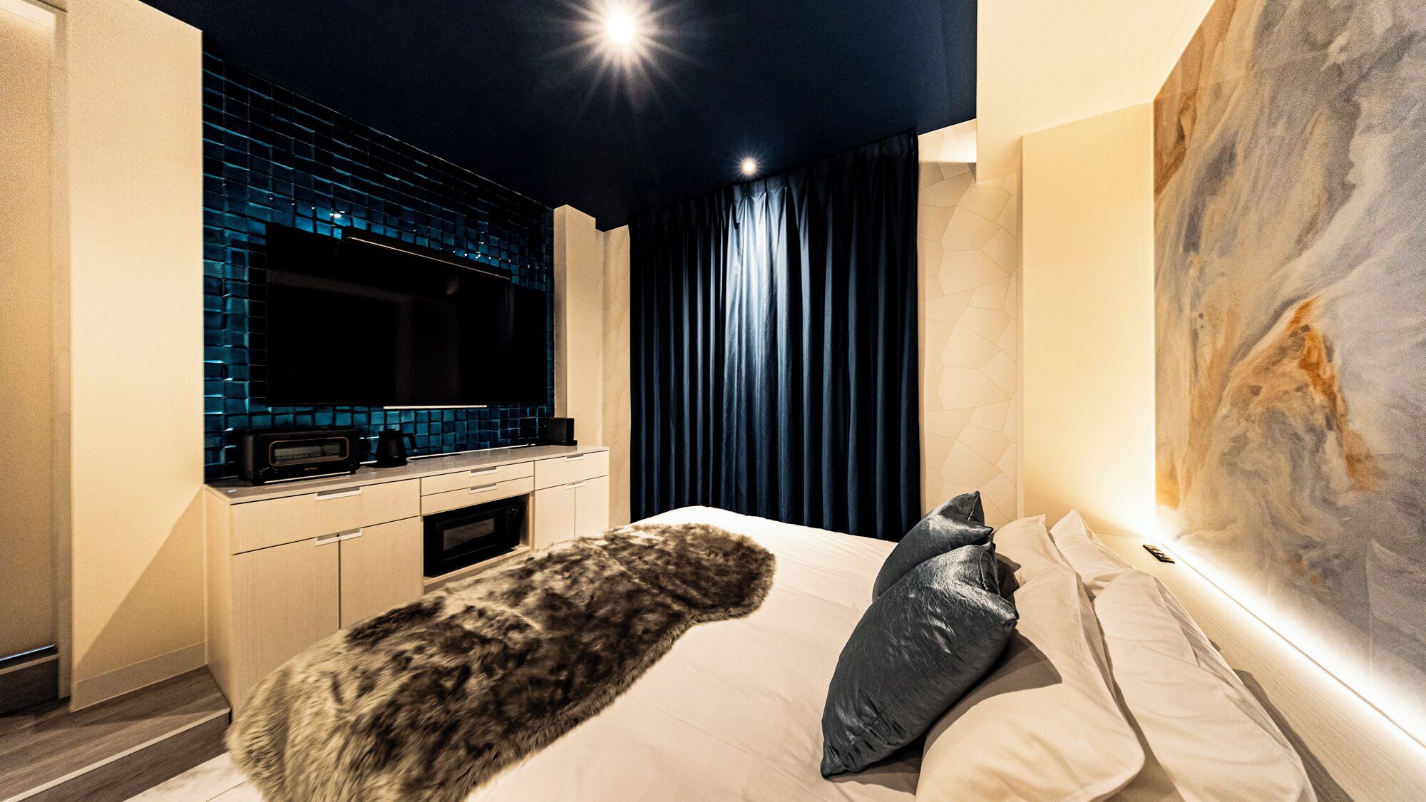 Quarto moderno com cama grande, manta de pele, pintura e área de cozinha com painel azul.