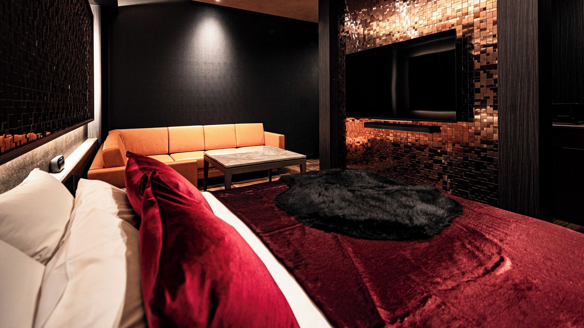 Quarto de hotel moderno com uma cama, um sofá, uma mesinha, uma televisão presa a uma parede de tijolos e um tapete de pele.