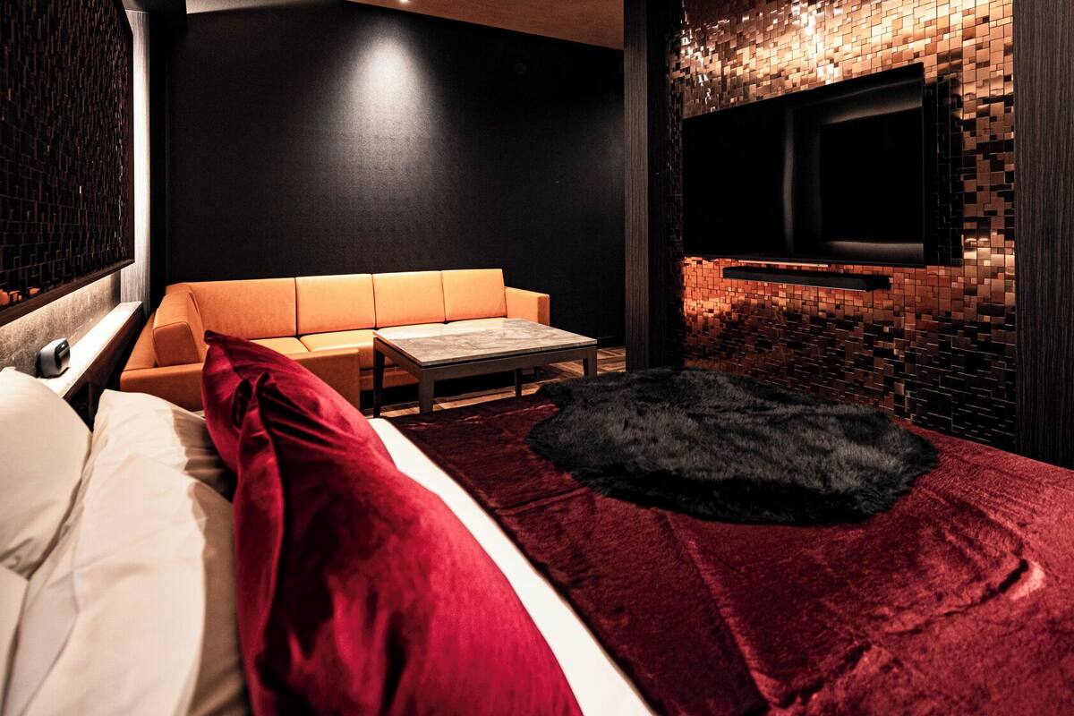 Habitación de hotel moderna con una cama, un sofá, una mesita y un televisor fijado a una pared de ladrillo, además de una alfombra de piel.