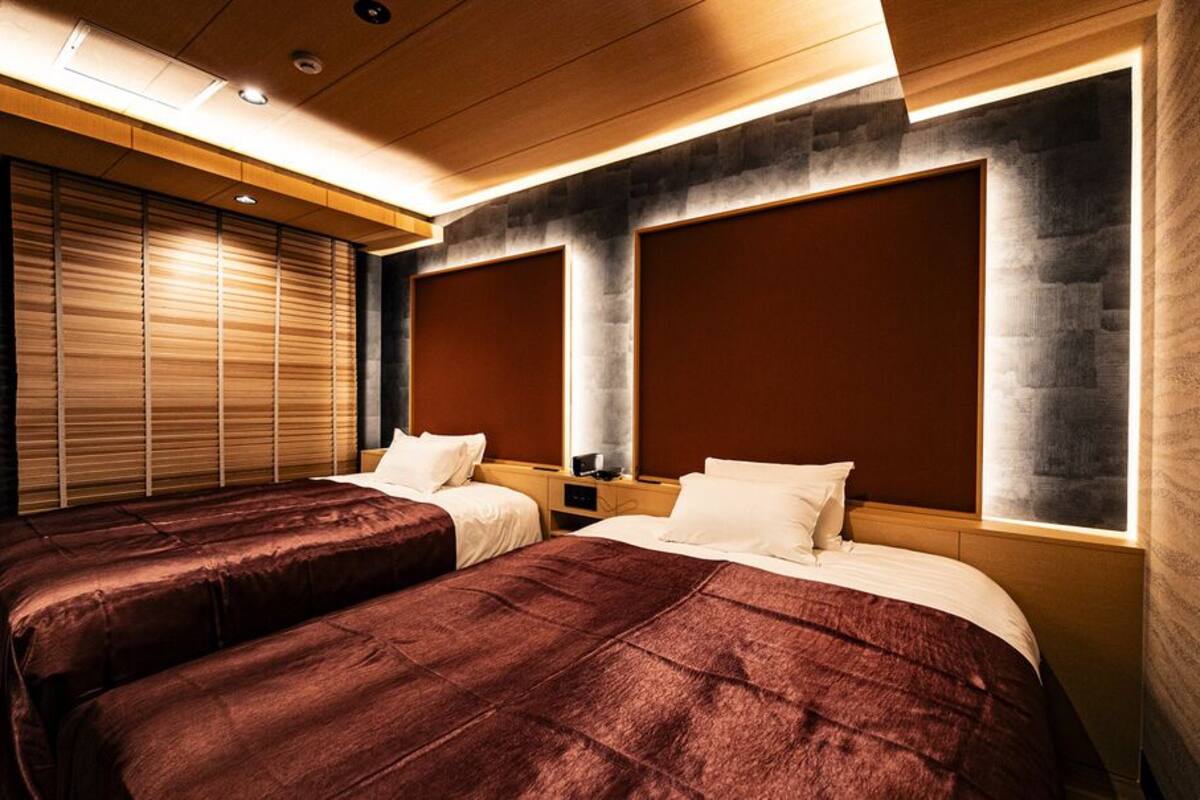 Habitación de hotel con dos camas, un ventanal grande con persianas y un diseño moderno que incorpora elementos de madera y piedra.