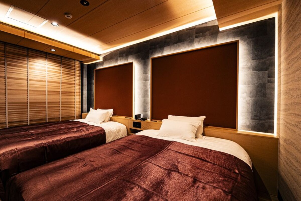 A hotel room with two beds, a large window with blinds, and a modern design with wooden and stone elements.