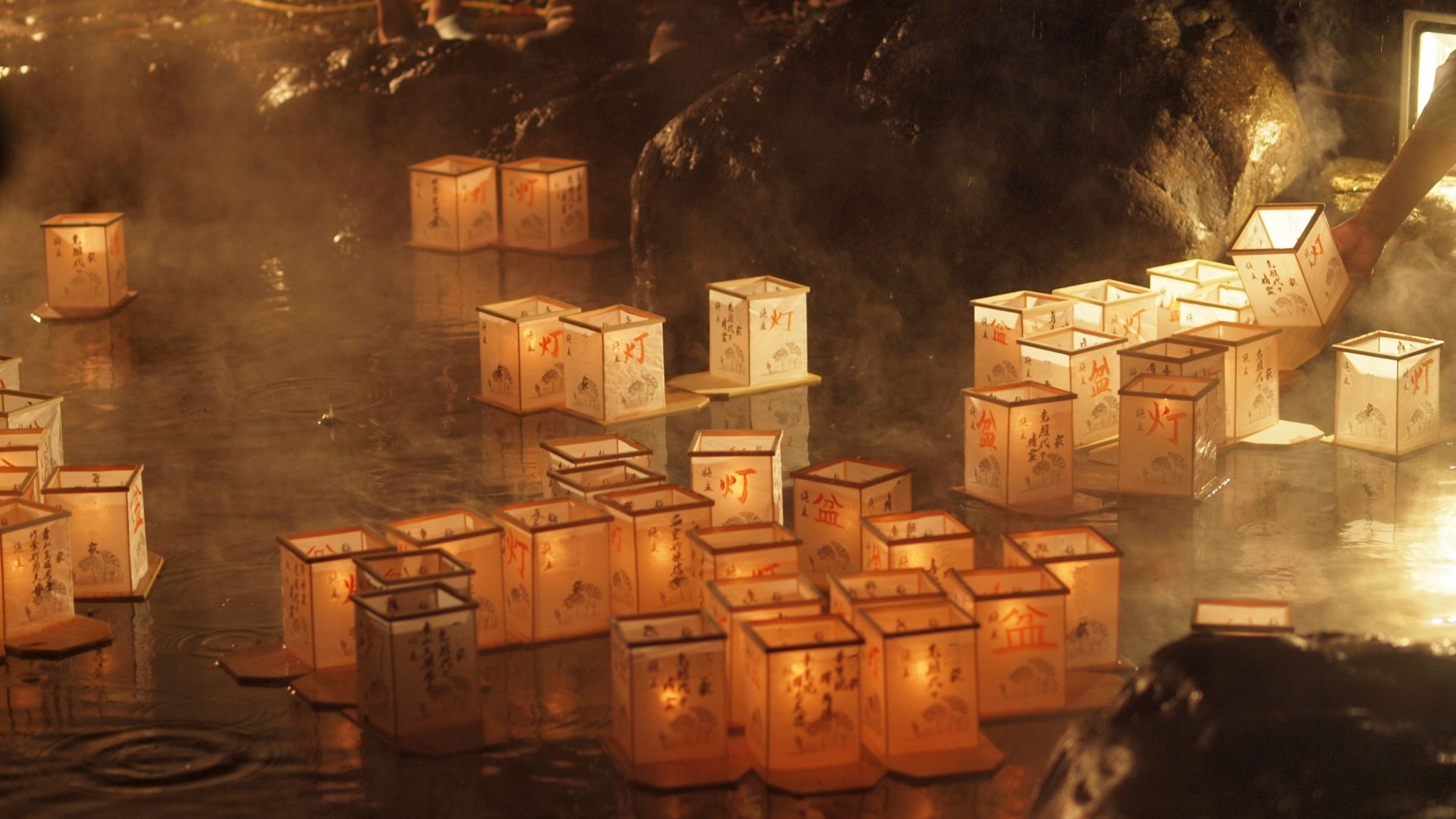 A series of illuminated lanterns floating on water, with a rocky background.
