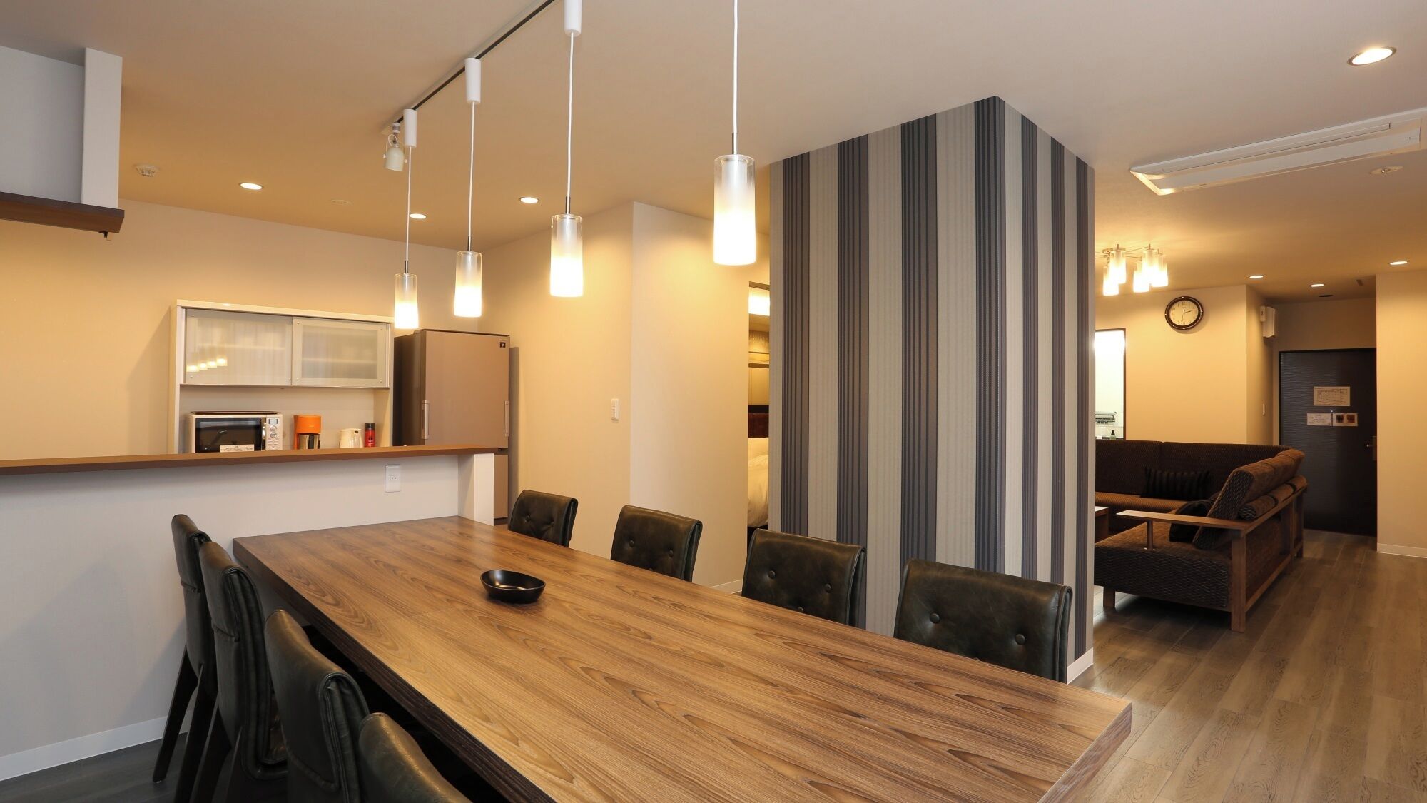 A modern hotel room with a wooden dining table, black leather chairs, a striped partition, and a sofa.