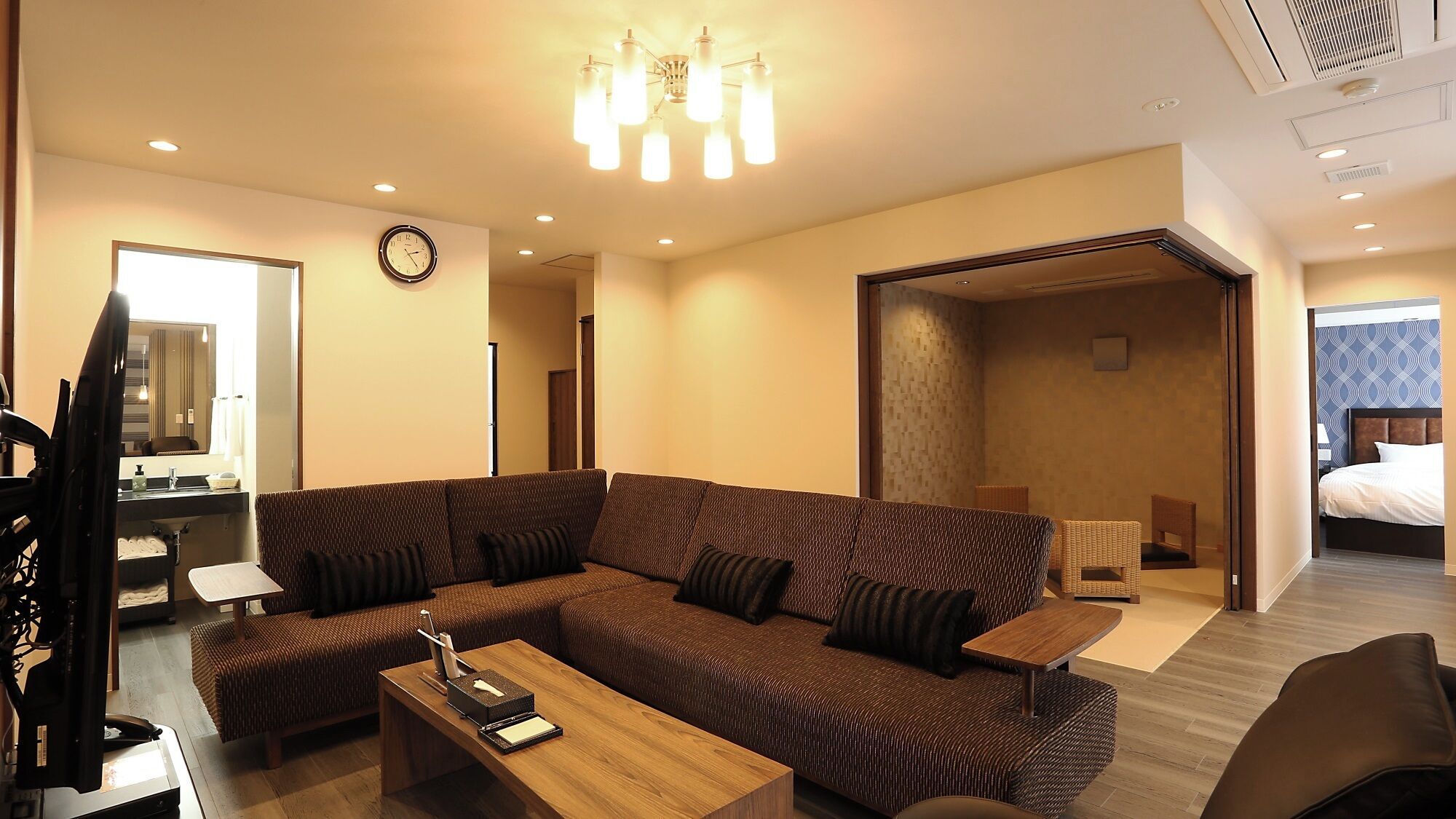 A modern hotel room with a large sectional sofa, a wooden coffee table, a television, and a bathroom visible through an open door.