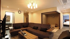 A modern hotel room with a large sectional sofa, a wooden coffee table, a television, and a bathroom visible through an open door. - Point Vacation Gero (Gero)