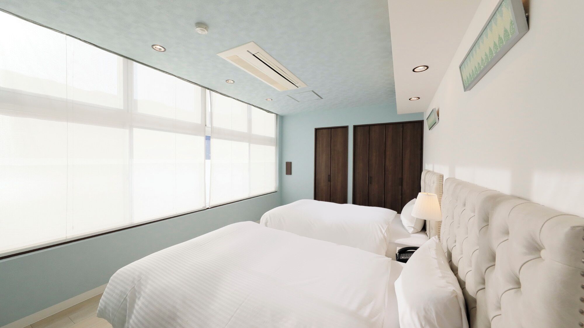 A hotel room with two beds, a large window, a wardrobe, and a wall-mounted lamp.