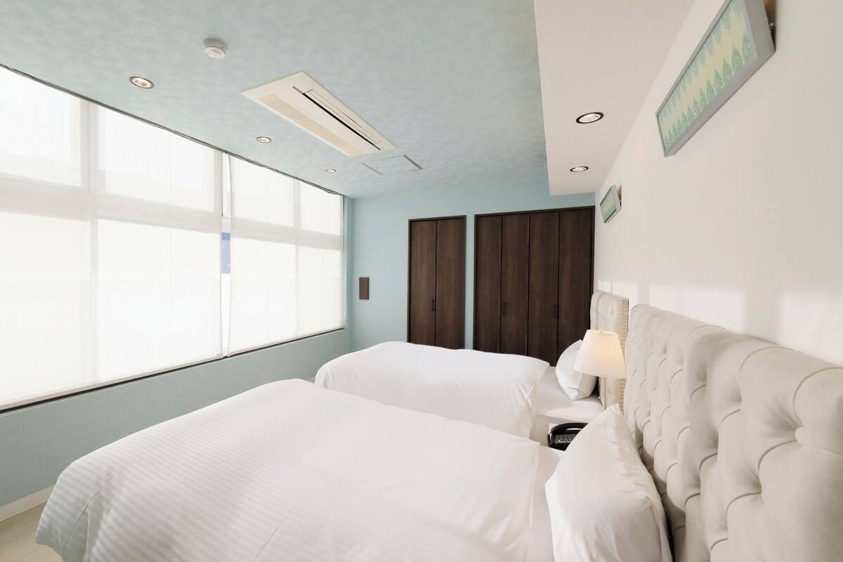 A hotel room with two beds, a large window, a wardrobe, and a wall-mounted lamp.