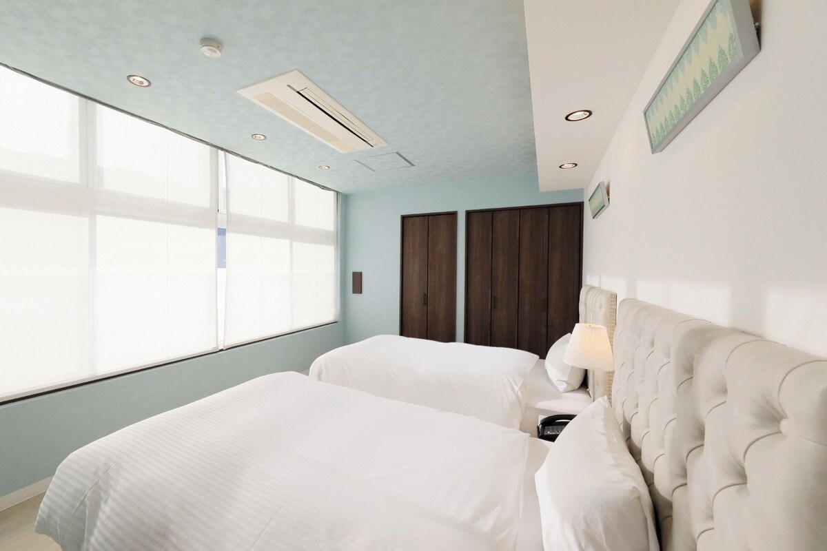A hotel room with two beds, a large window, a wardrobe, and a wall-mounted lamp.