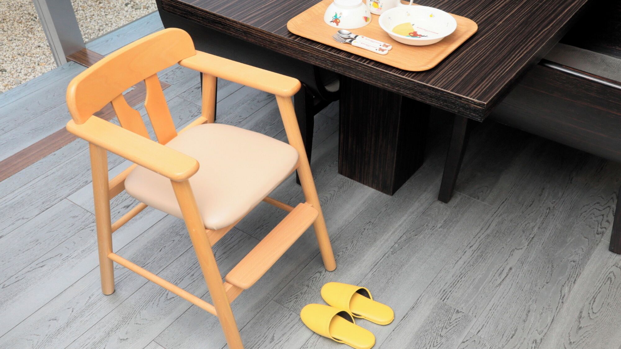 Una silla de madera con cojín, una mesa de comedor con una bandeja de platos y cubiertos, y un par de zapatillas amarillas en el suelo.