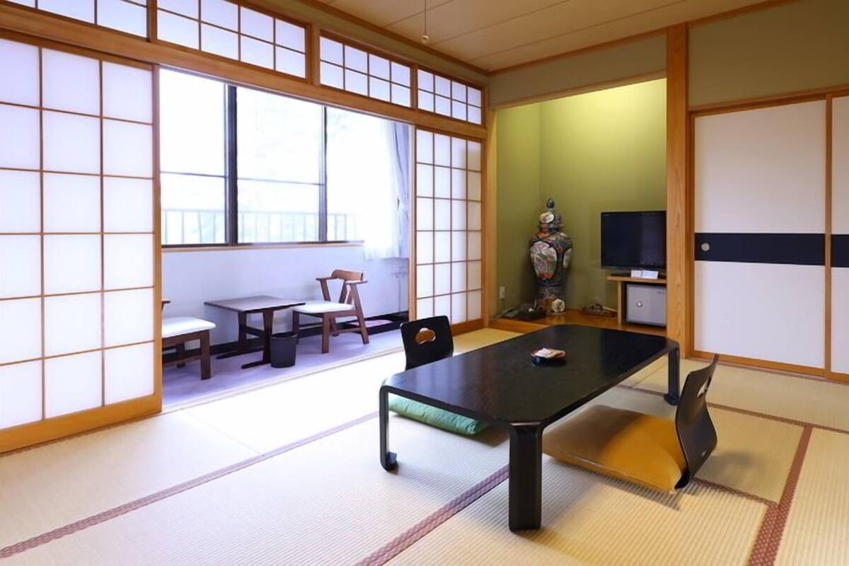Ein traditioneller japanischer Raum mit Tatami-Boden, einem niedrigen Tisch, Stühlen, einem Fernseher und einer Dekorvase.