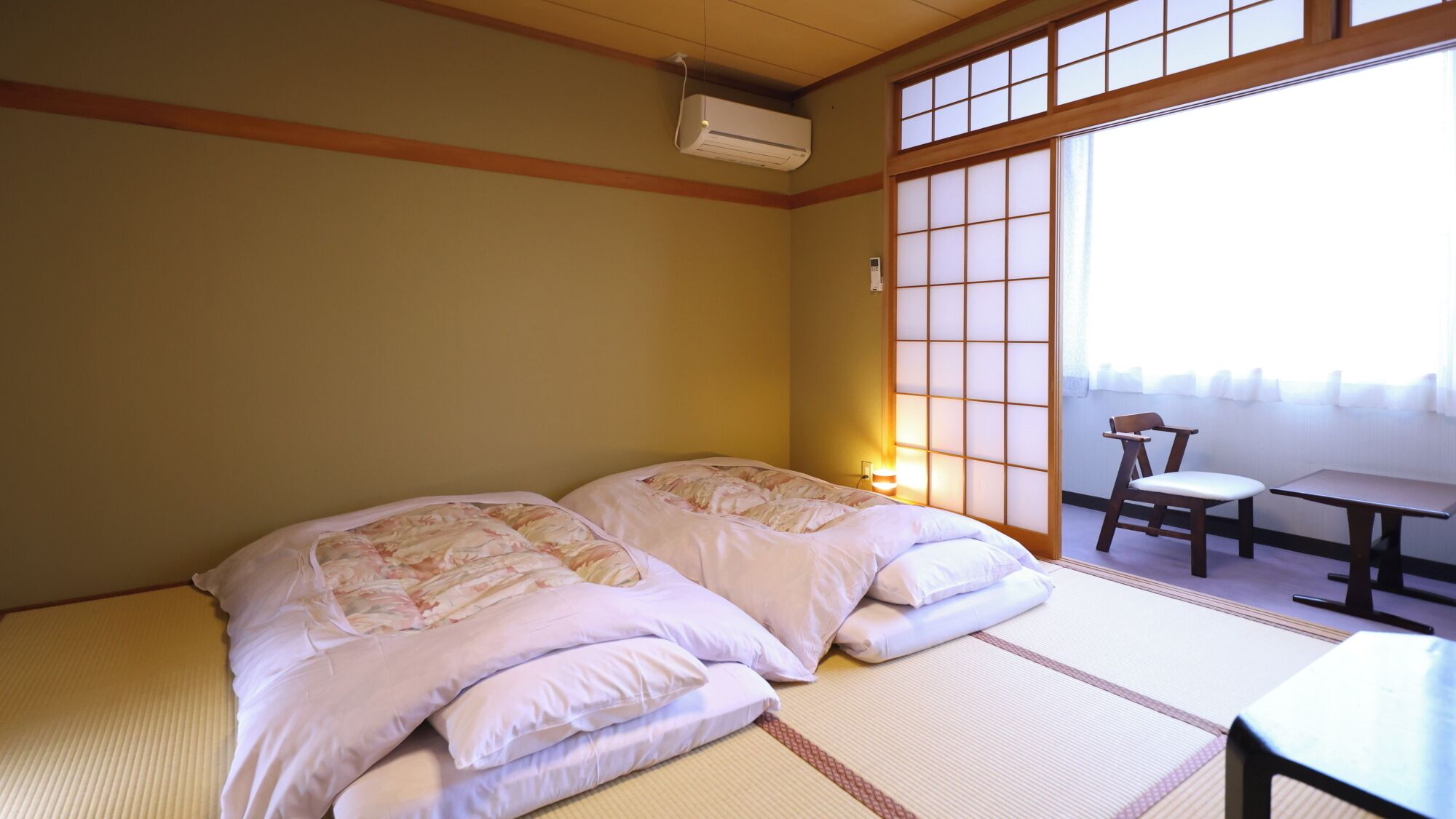 A traditional Japanese-style room with tatami flooring, two beds with floral bedding, a sliding shoji screen, and a small table with a chair.