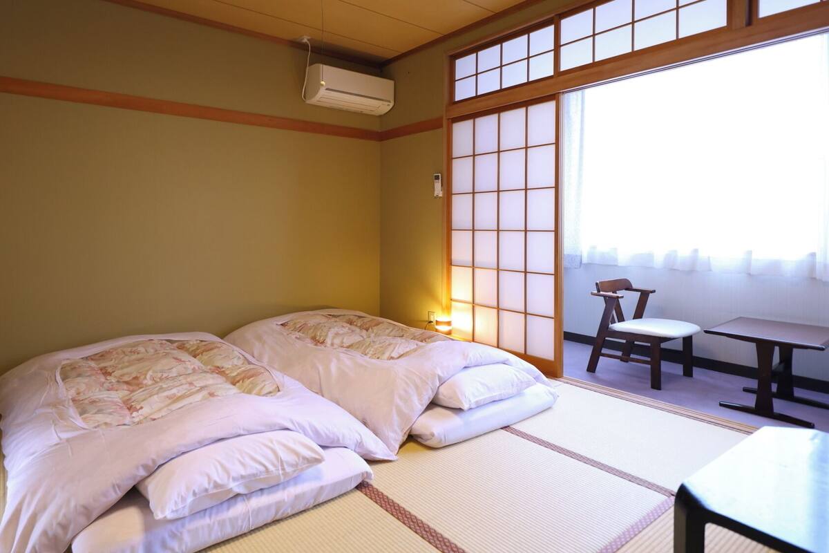Ein traditioneller japanischer Raum mit Tatami-Bodenbelag, zwei Betten mit floralen Betttextilien, einem verschiebbaren Shoji-Bildschirm und einem kleinen Tisch mit Stuhl.