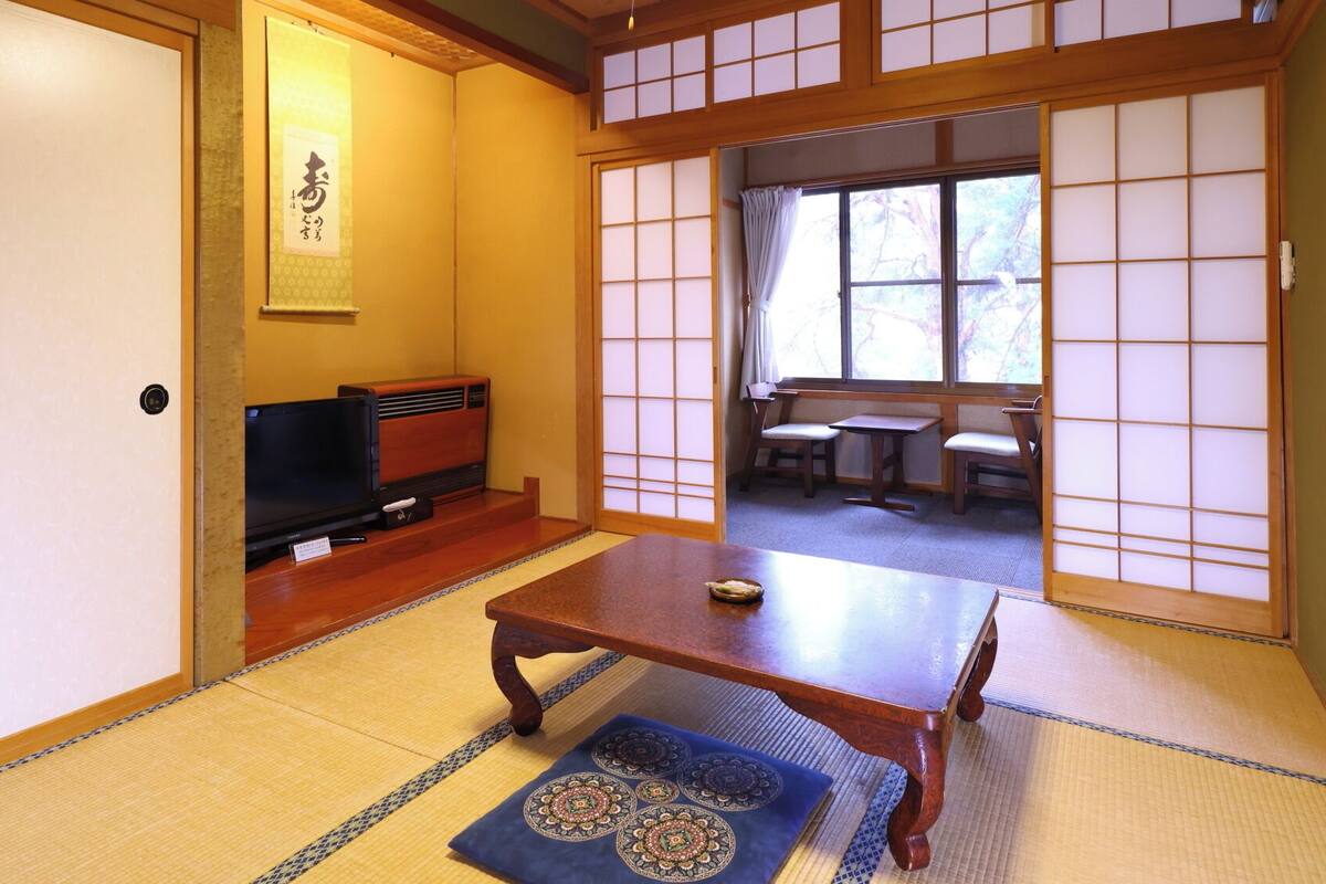 Ein traditioneller japanischer Raum mit Tatami-Böden, einem niedrigen Tisch, einem Fernseher und Schiebetüren zu einem Essbereich.