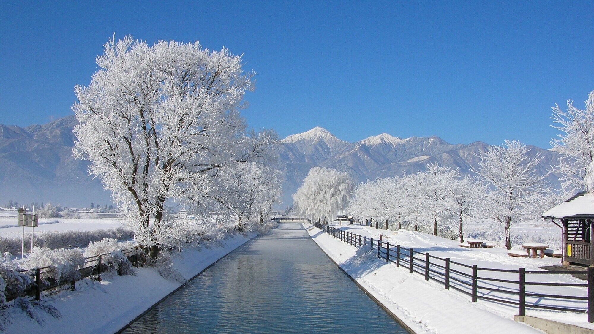 Une rivière recouverte de neige, bordée d’une clôture en bois et de bancs, et entourée d’arbres et de montagnes.