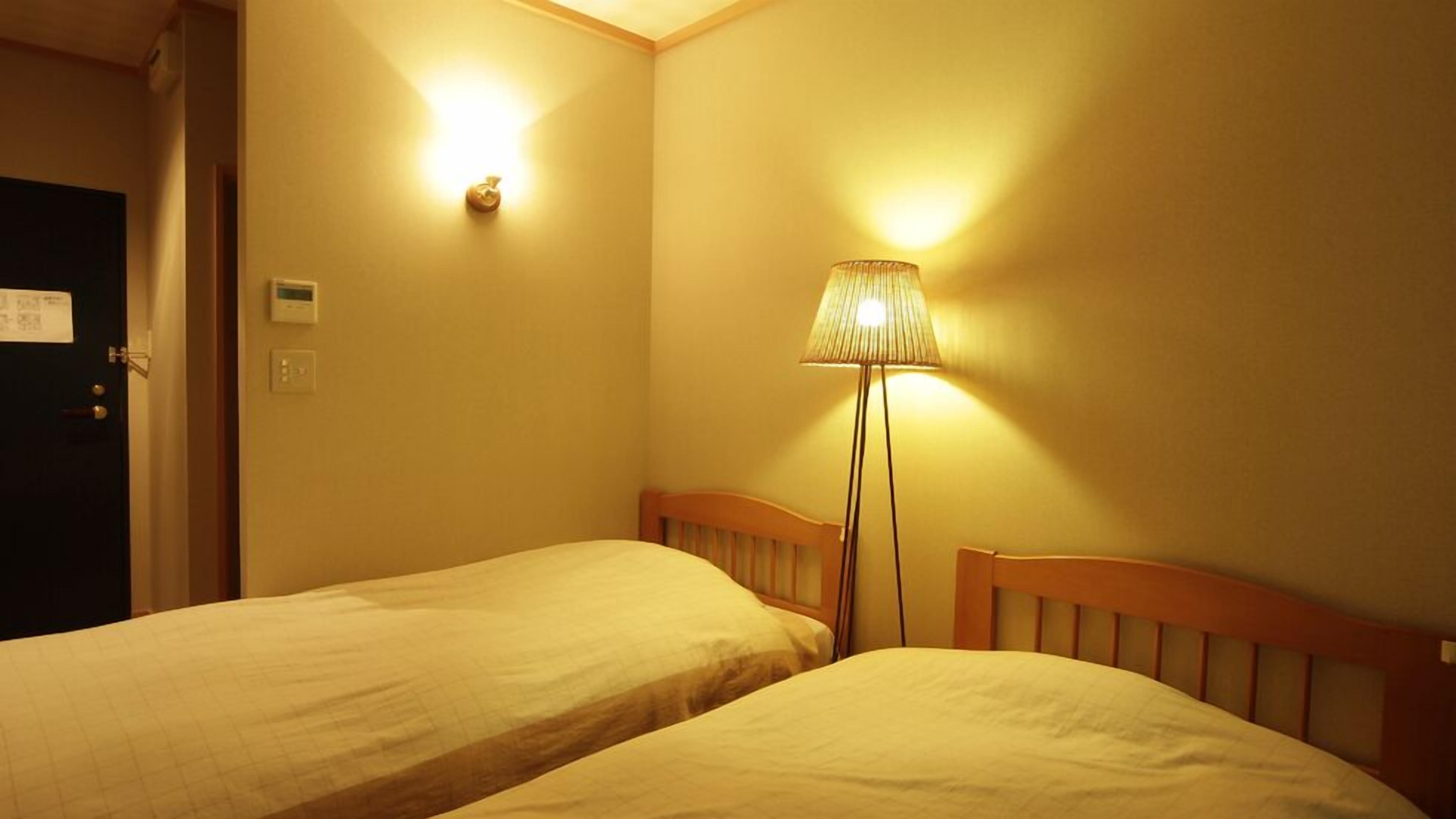 Habitación de hotel con dos camas individuales, una mesita de noche, una lámpara y una puerta.