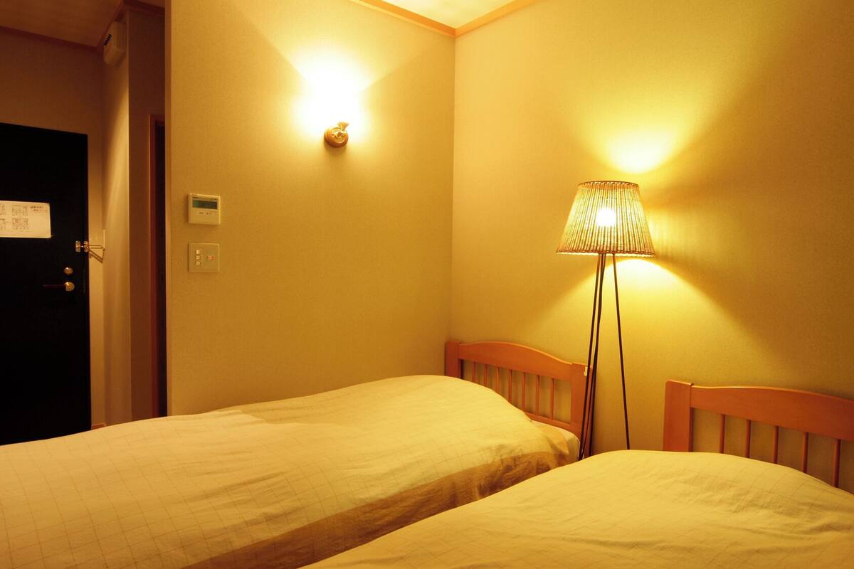A hotel room with two single beds, a standing lamp, a door, and a wall-mounted light fixture.