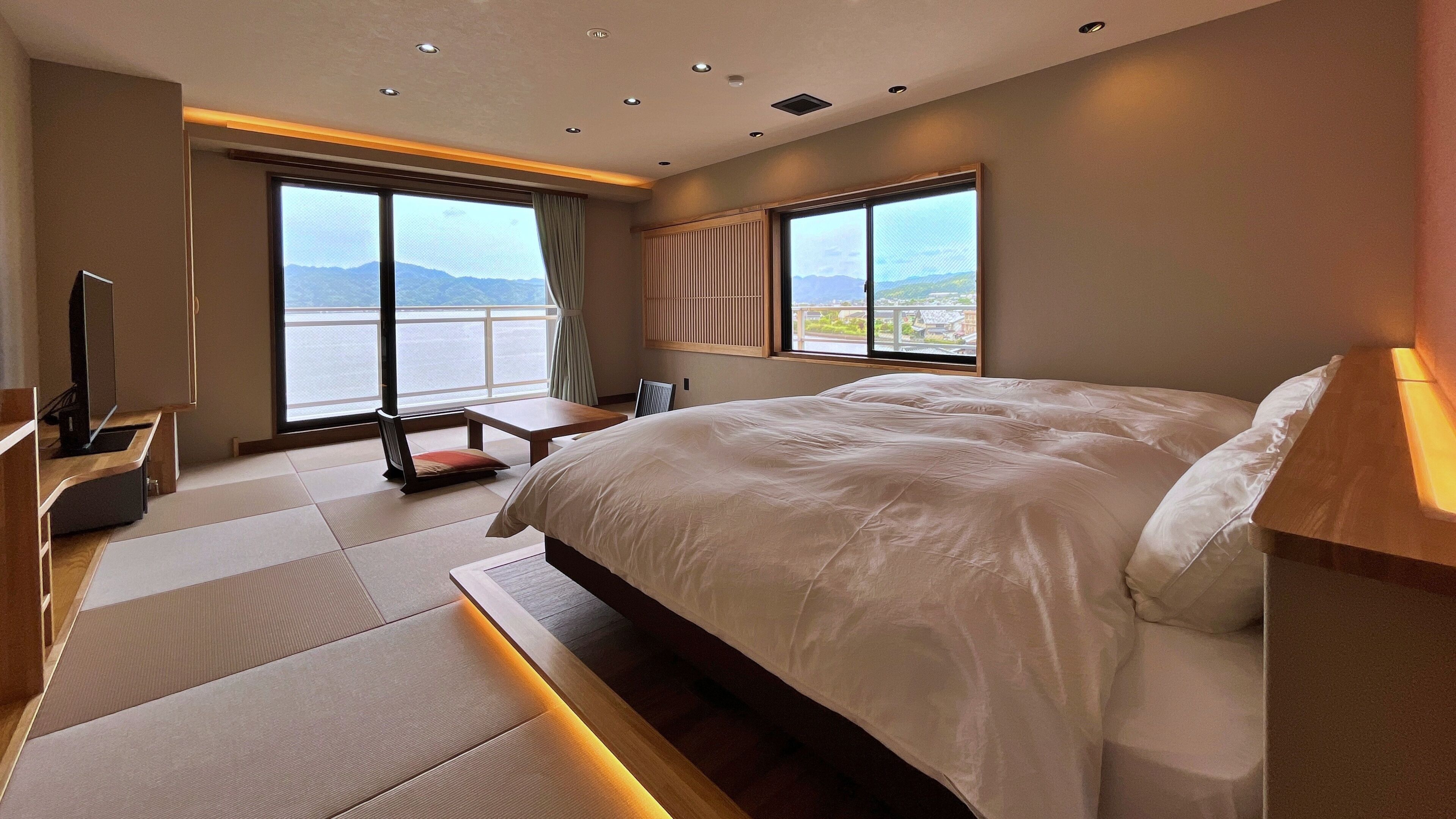 A modern hotel room with a large bed, a TV, a desk, and a view of the mountains.