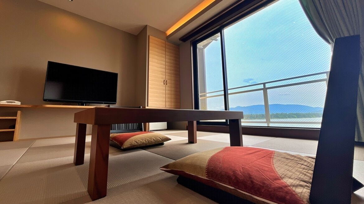A modern hotel room with tatami flooring, a television, a large window with a view of mountains, and a wooden bench.
