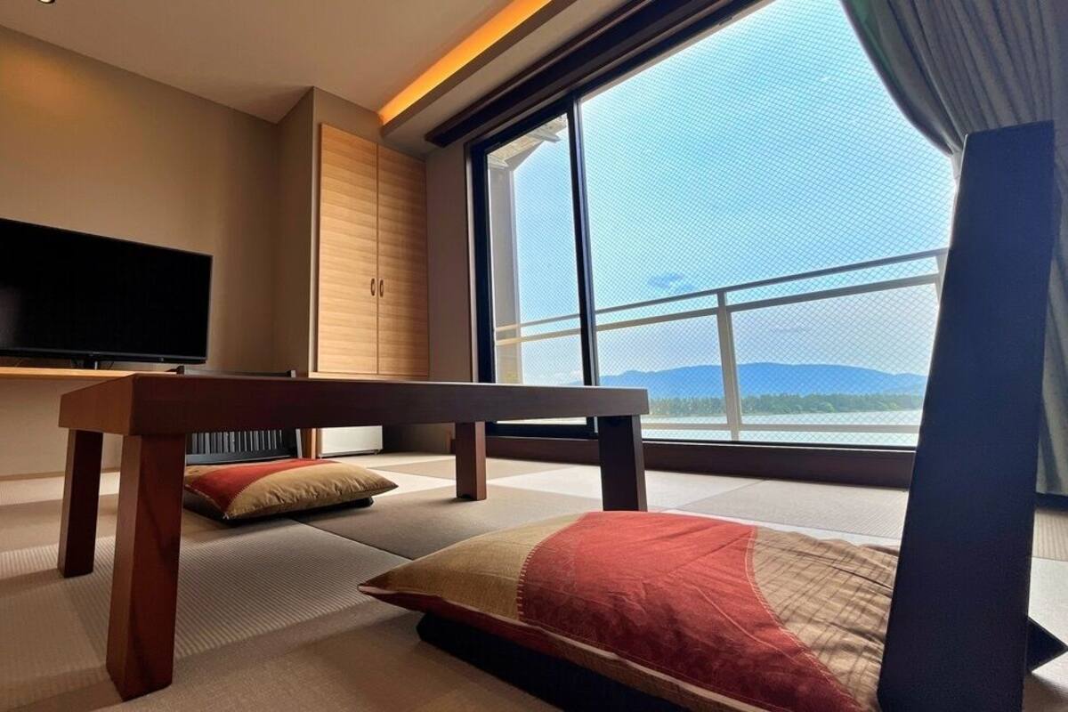 A modern hotel room with tatami flooring, a television, a large window with a view of mountains, and a wooden bench.