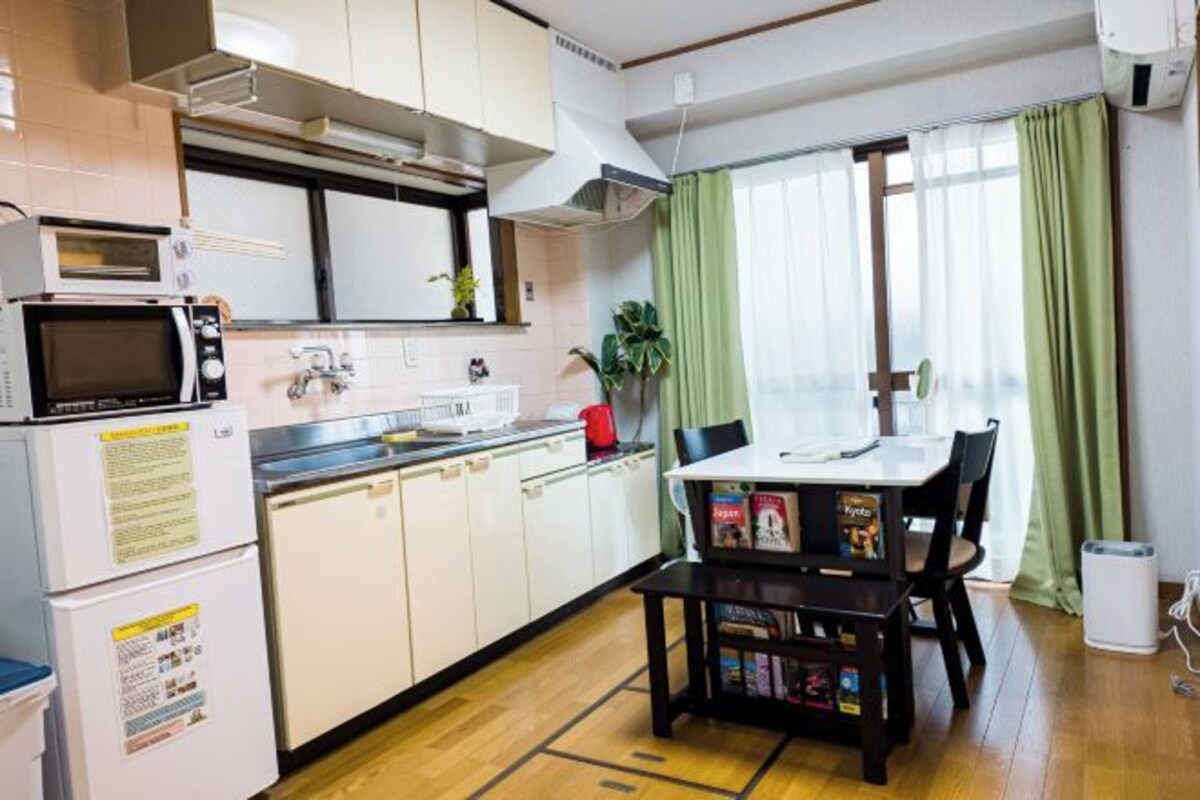 Ett kompakt kök med mikrovågsugn, diskho och skåp. Där finns ett matbord med stolar och en liten krukväxt.