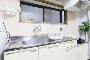 A kitchen with a stainless steel sink, white cabinets, a window above the sink, and a plant on the right. - Comodita Takeyacho 1f (Kyoto)