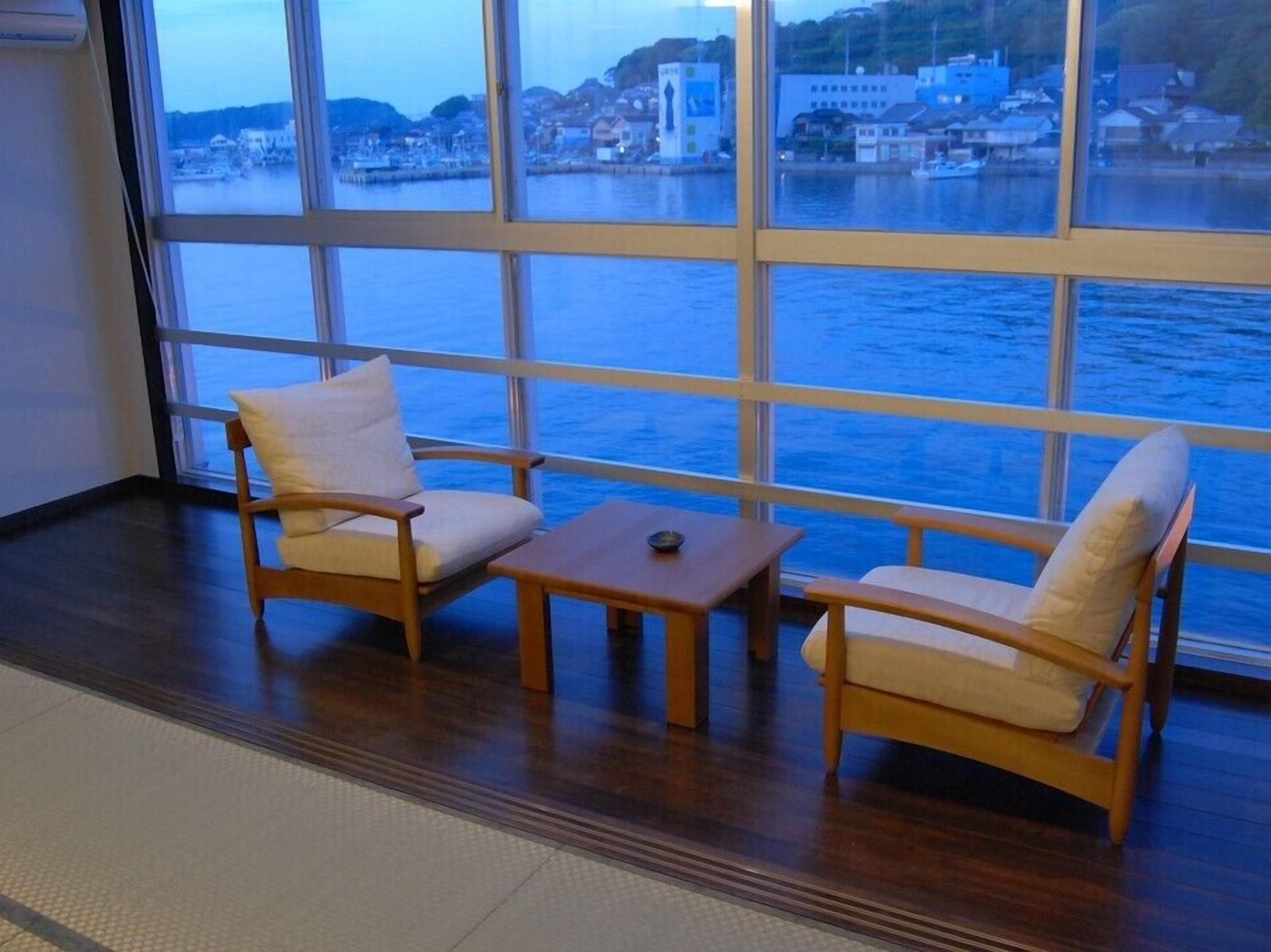 Quarto com uma janela ampla que dá para a água, duas cadeiras de madeira, uma mesa pequena e piso de azulejos.