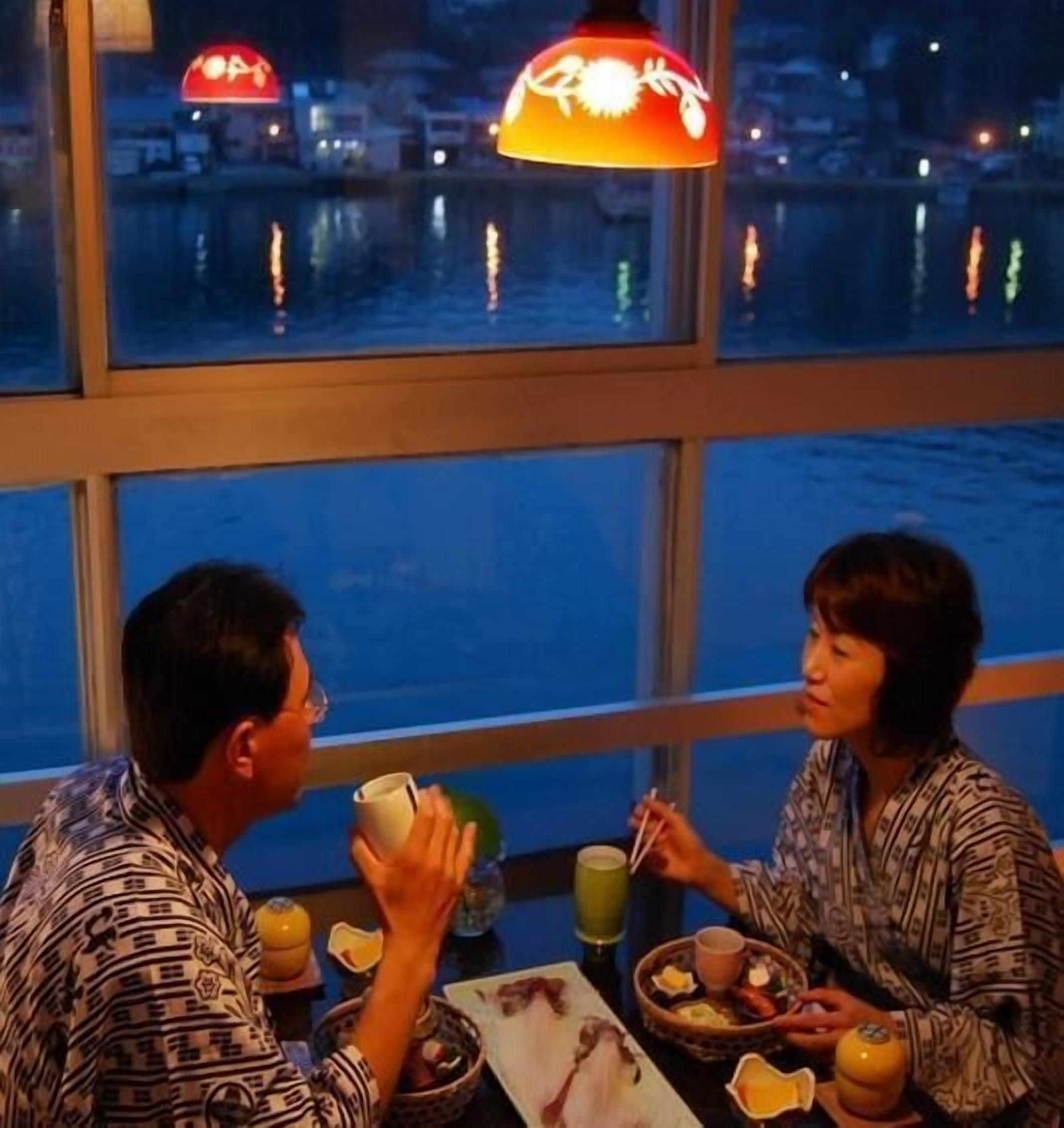 A dining area with a view of a body of water, a table with food and drinks, and a hanging lamp.