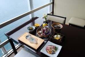 Uma mesa de jantar com diversos pratos, um vaso de flores e vista para o mar através de grandes janelas.