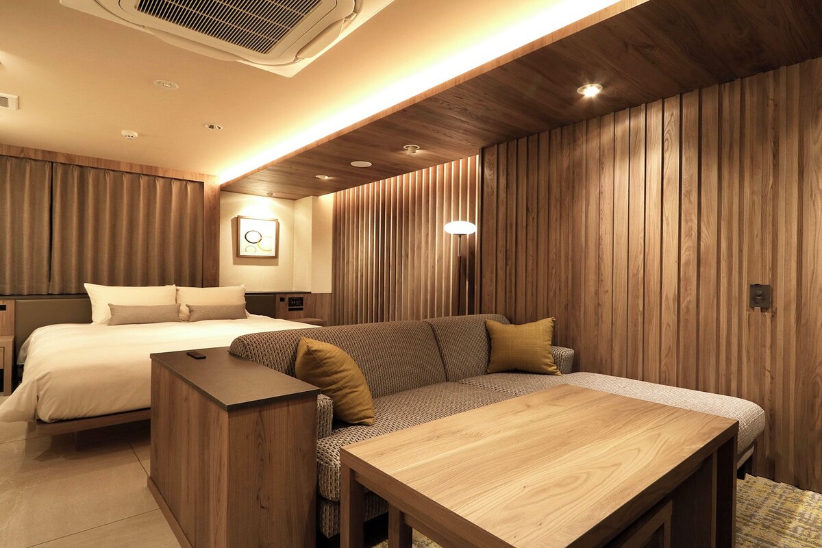 A hotel room with a bed, a sofa, a wooden table, and a wall-mounted lamp.