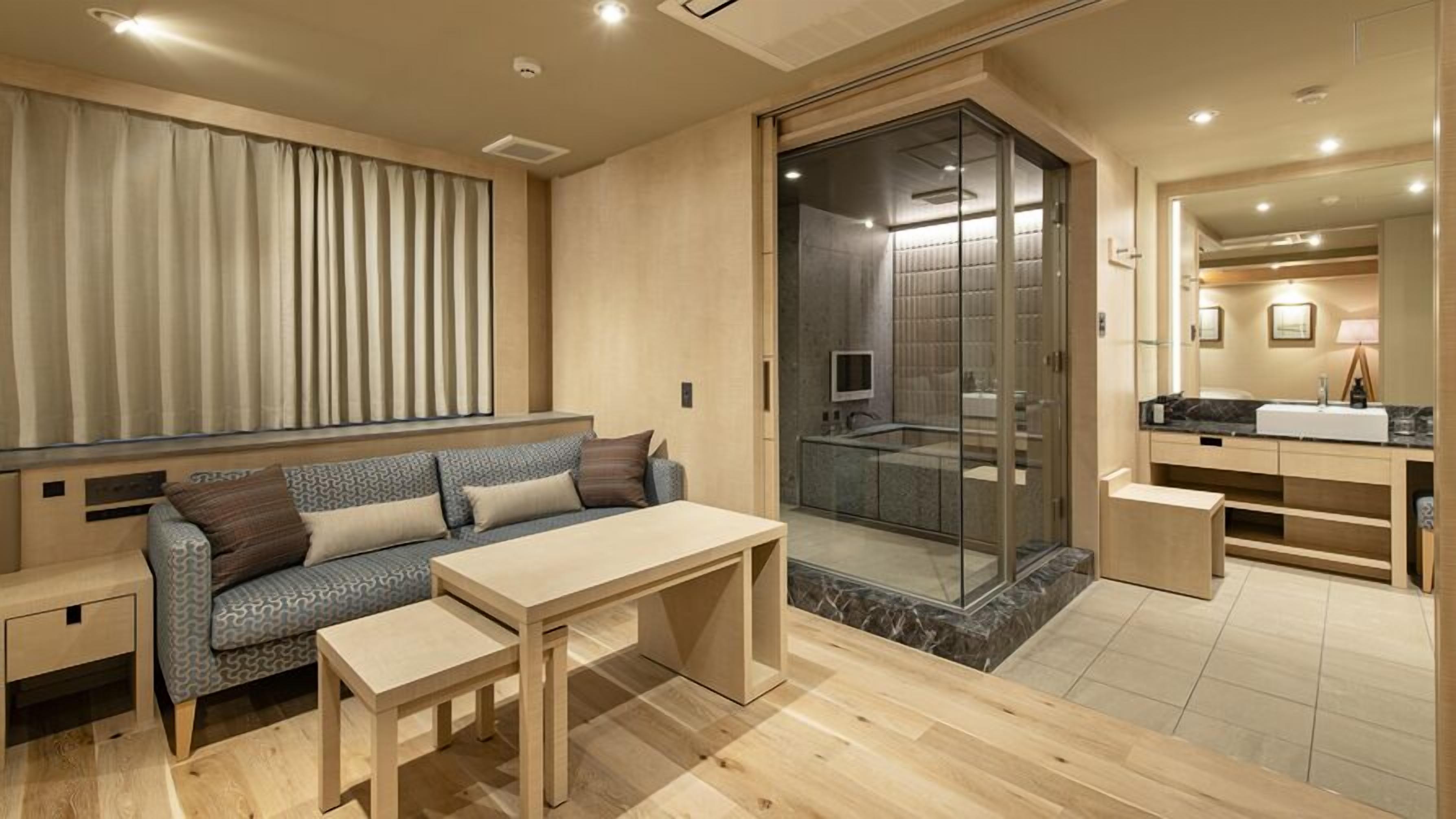 A modern hotel room with a sofa, a small table, a glass-enclosed shower, a vanity with a mirror, and a window with curtains.