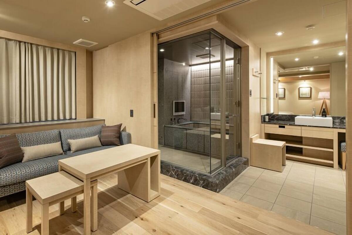 Ett modernt hotellrum med en soffa, ett litet bord, en dusch med glasväggar, en sminkbord med spegel och ett fönster med gardiner.