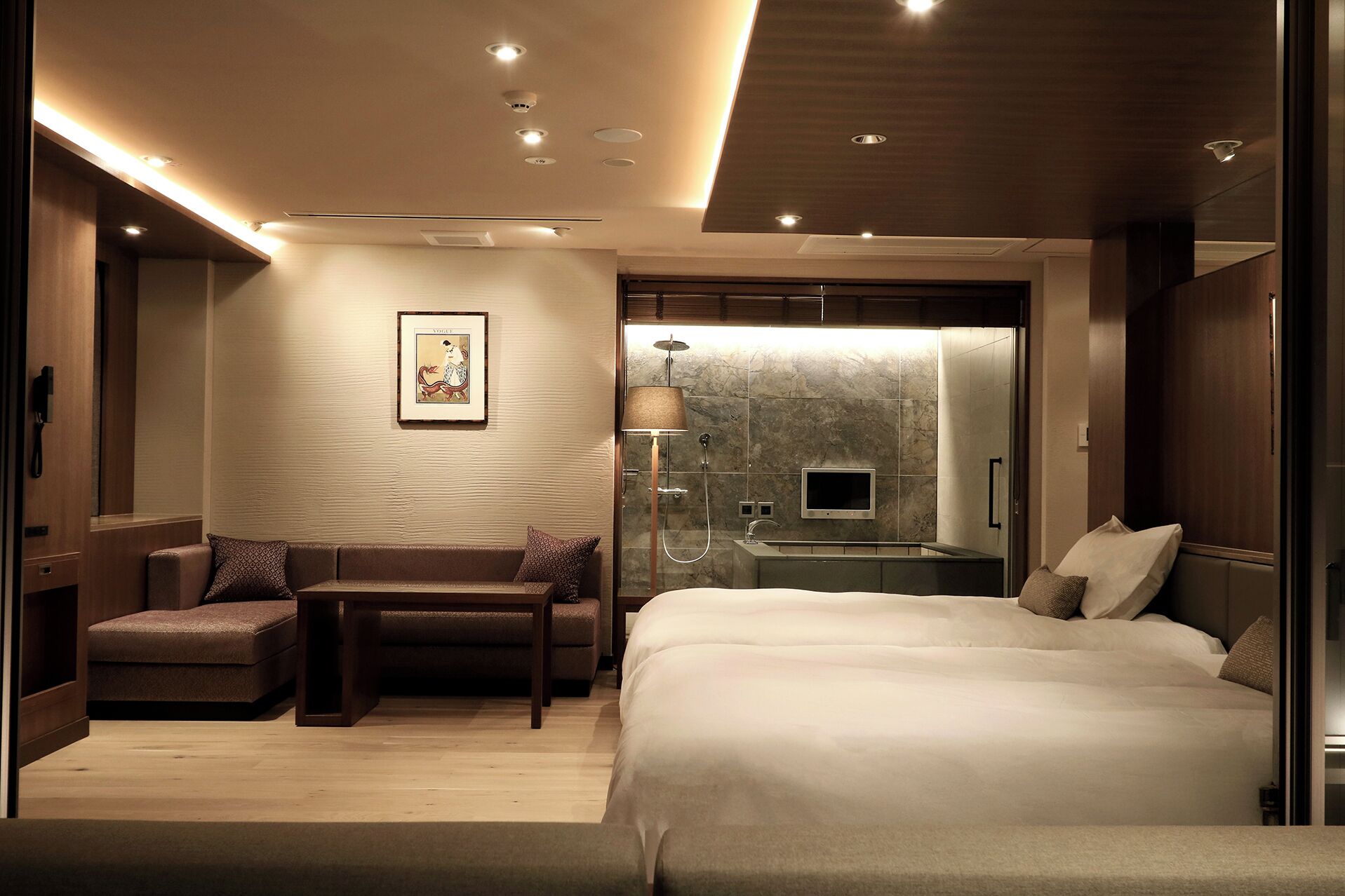 A modern hotel room with a large bed, a seating area with a table, a shower area, and a mounted television.