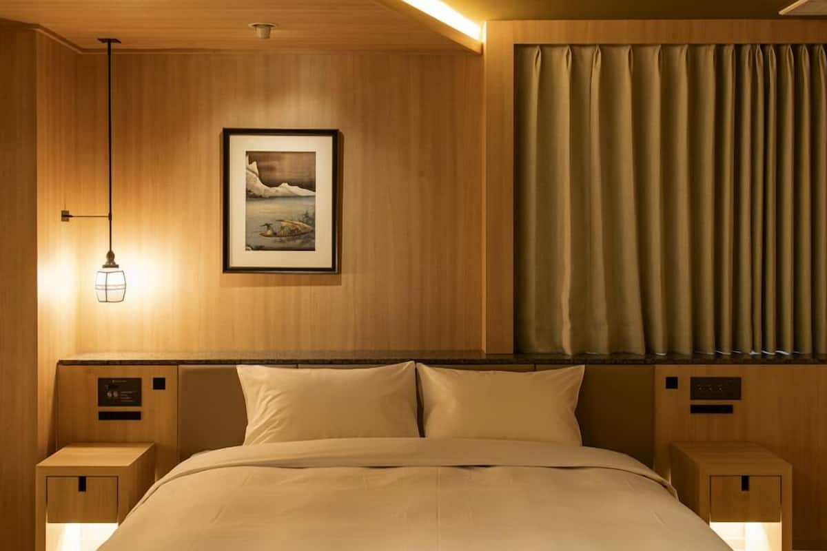 Ett hotellrum med en sänggavel i trä, en inramad tavla, en säng med vita sängkläder och ett fönster med gardiner.