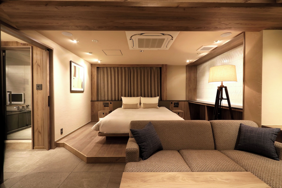 A modern hotel room with a bed, a sofa, a desk, and a bathroom visible through an open door.