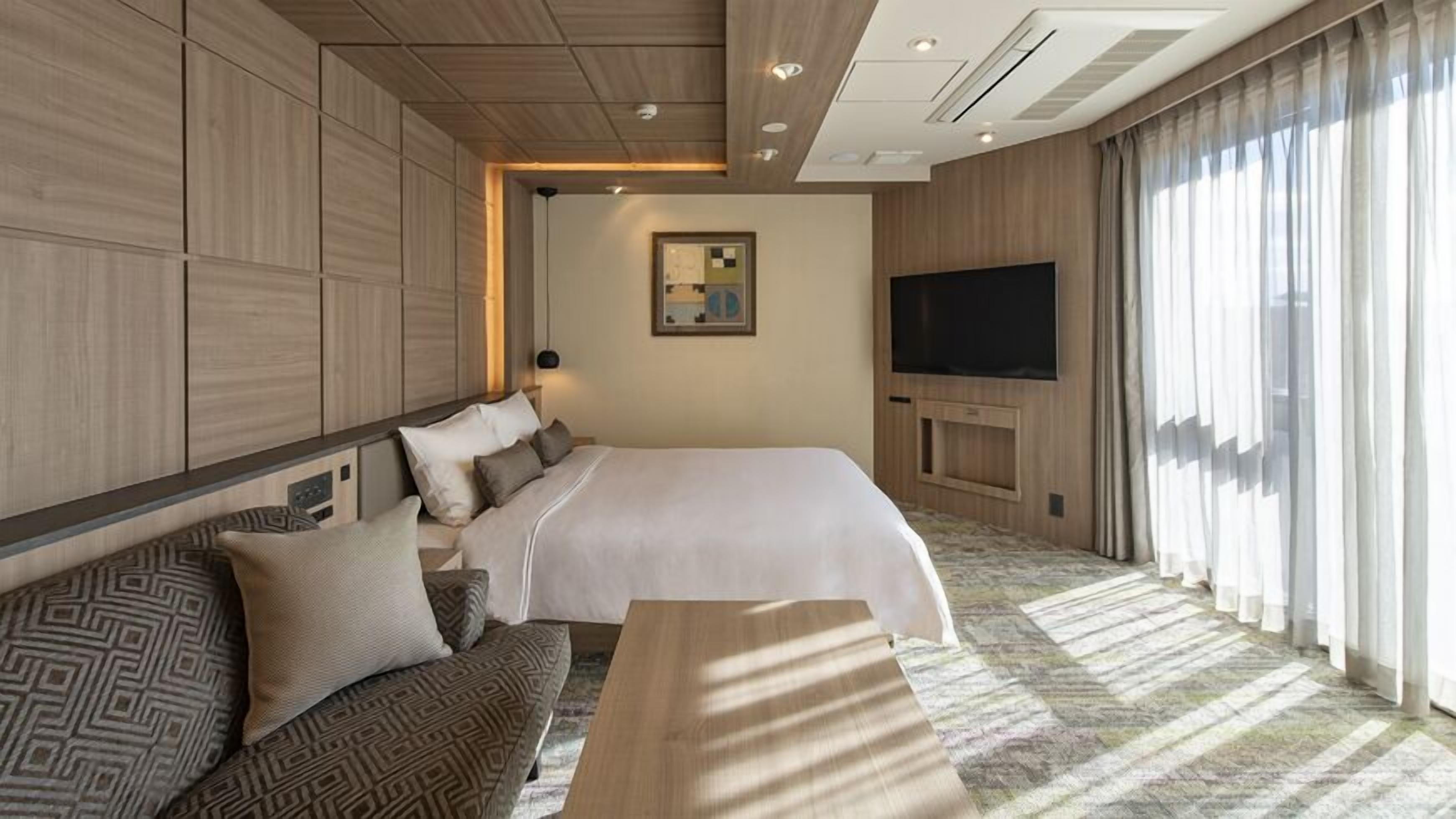 A hotel room with a large bed, a sofa, a TV, and a large window with curtains.