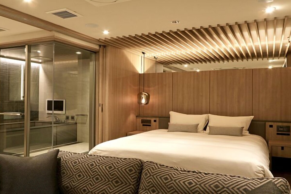 A modern bedroom with a large bed, a glass-enclosed shower, and a wooden ceiling design.