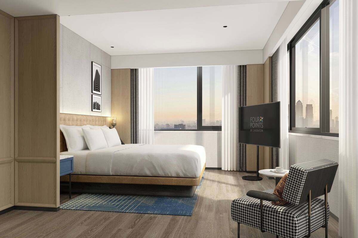 A modern hotel room with a large bed, a TV, a chair, and a view of the city.