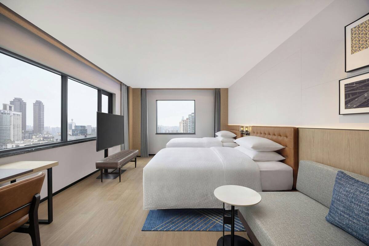 A hotel room with a large window, a bed, a sofa, a desk, and a TV.
