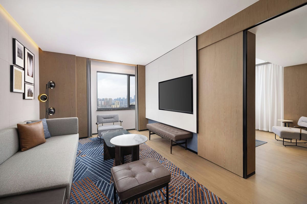 A modern hotel room with a sofa, a round table, a bench, a TV, and a city view.