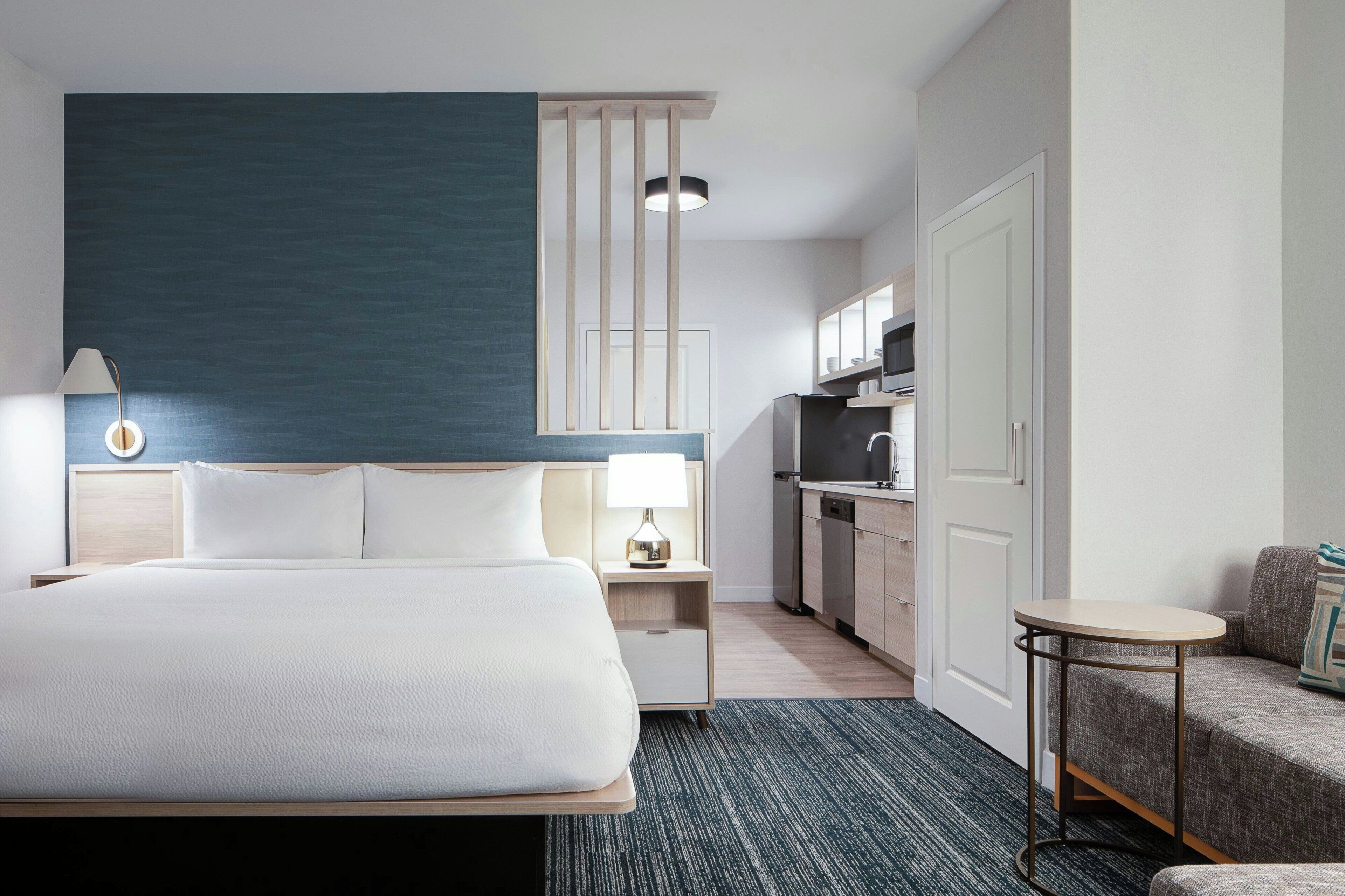 A hotel room with a bed, bedside table, lamp, and a kitchenette area.
