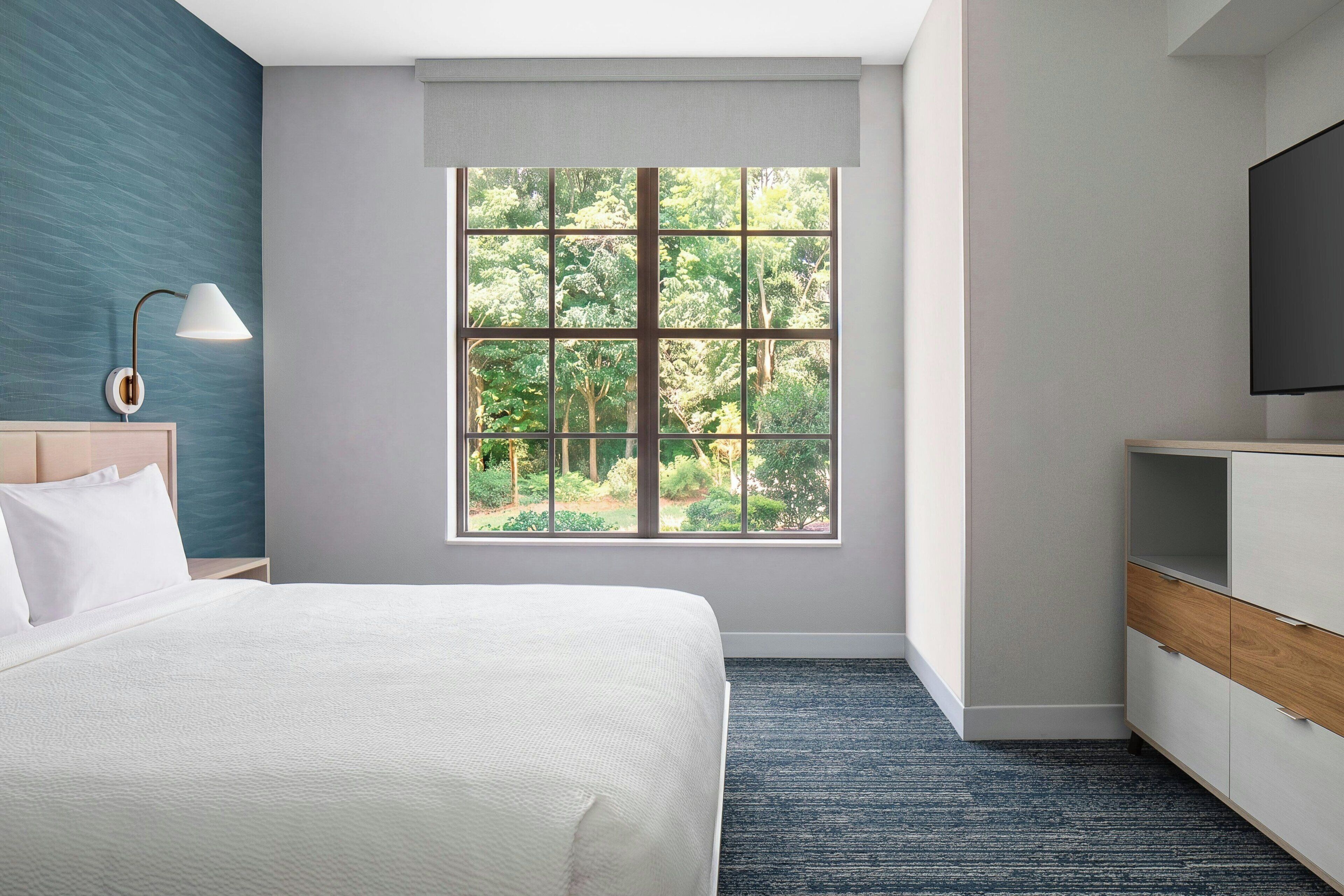 A hotel room with a bed, a desk, a chair, a TV, and a window with a view of trees.